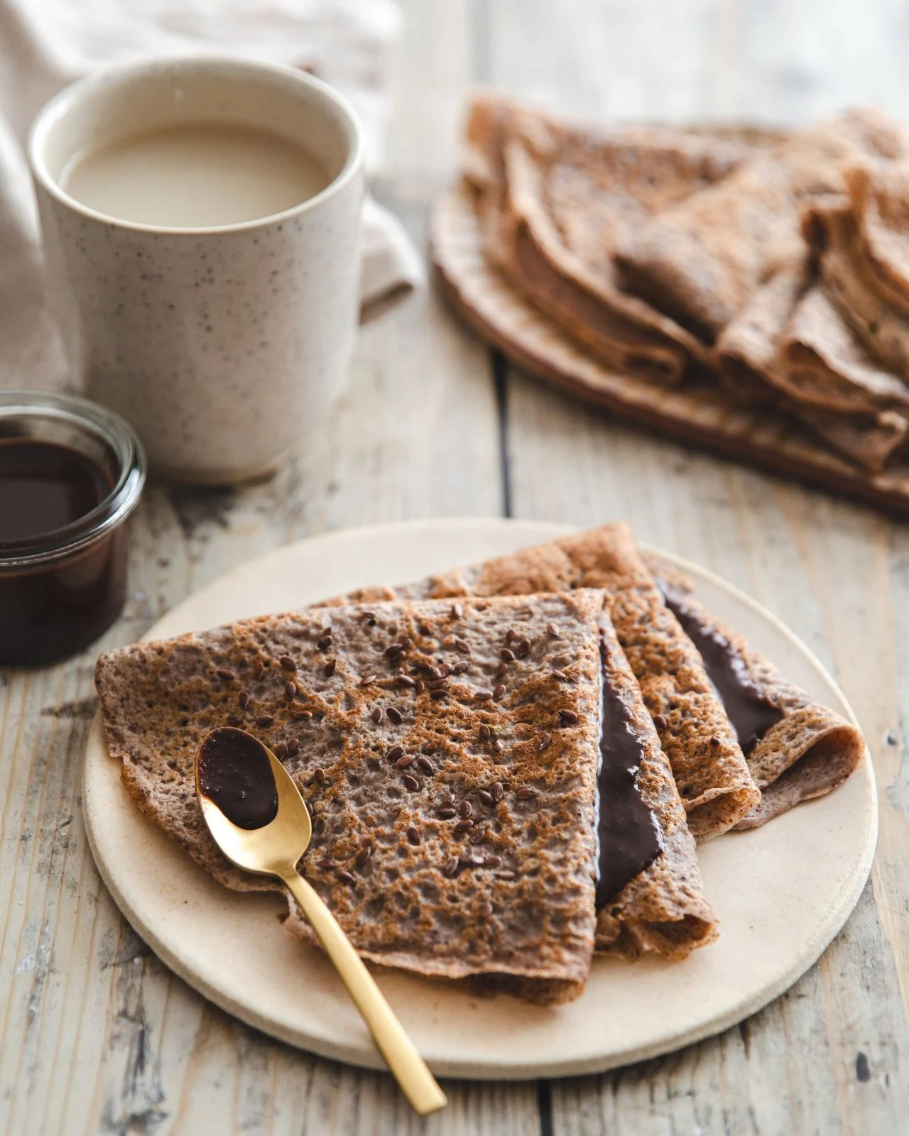 Flax flour crêpes folded on a plate, chocolate spread, flaxseed and a bowl of coffee